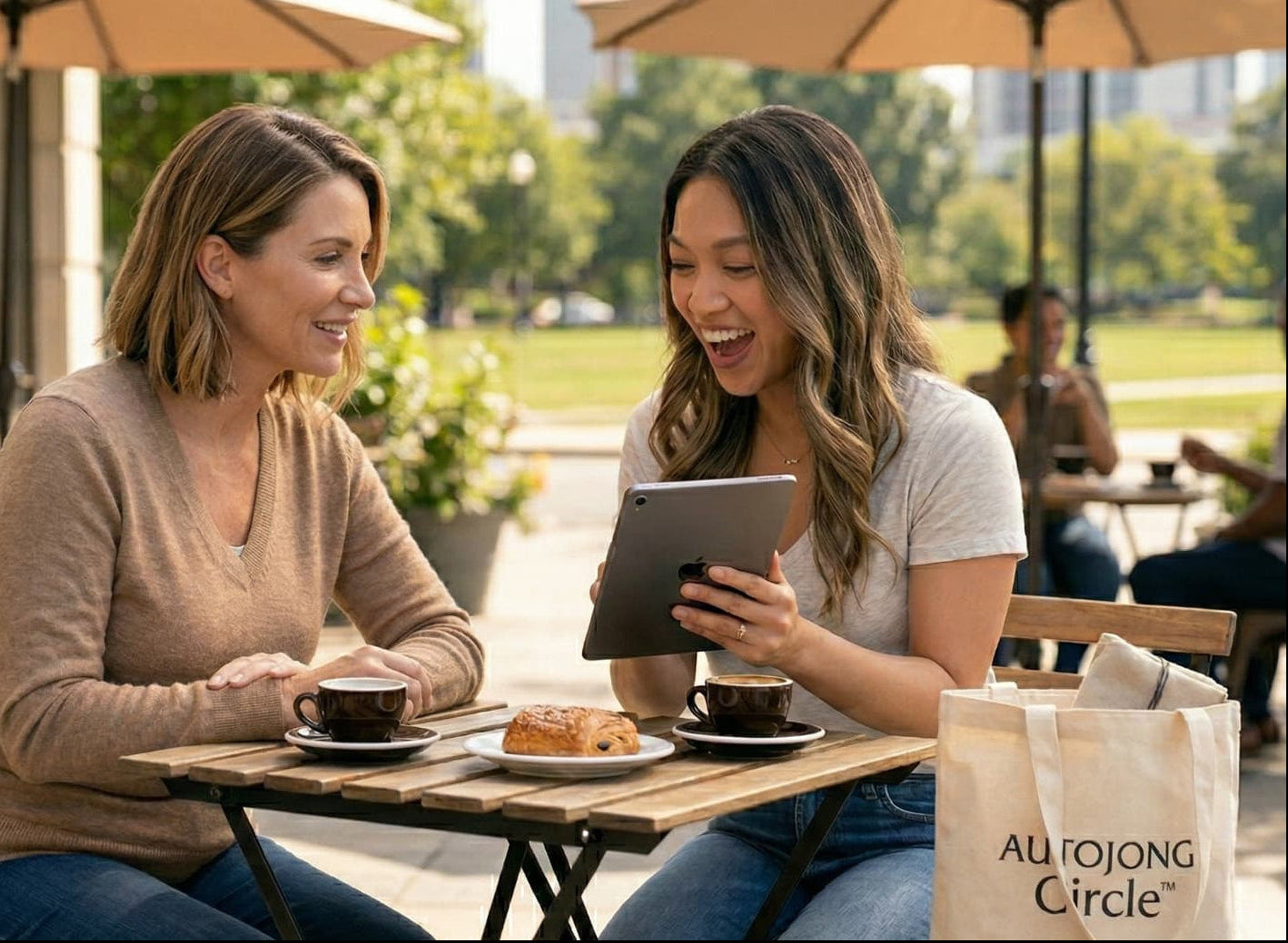 Two women enjoying coffee outdoors, smiling while sharing something on a tablet — a natural, relaxed moment of connection and trust